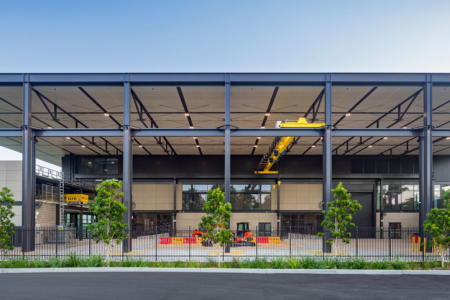 Front view of an industrial-style TAFE training facility with exposed steel frame, yellow overhead crane and young trees.