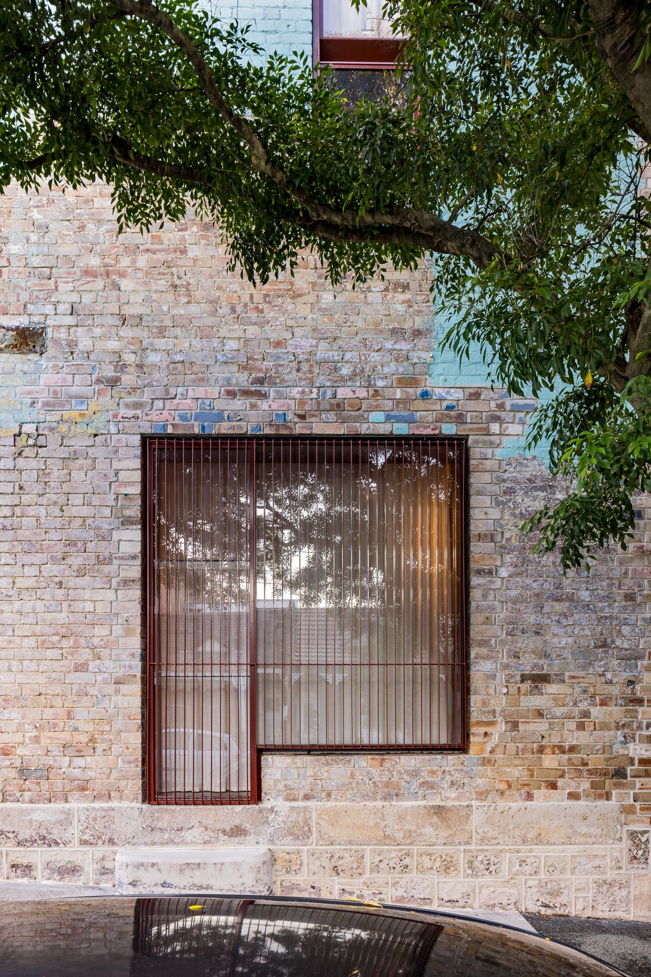 Weathered heritage brick wall with a square window framed in rust-red steel and fitted with vertical slatted screens under a leafy tree.