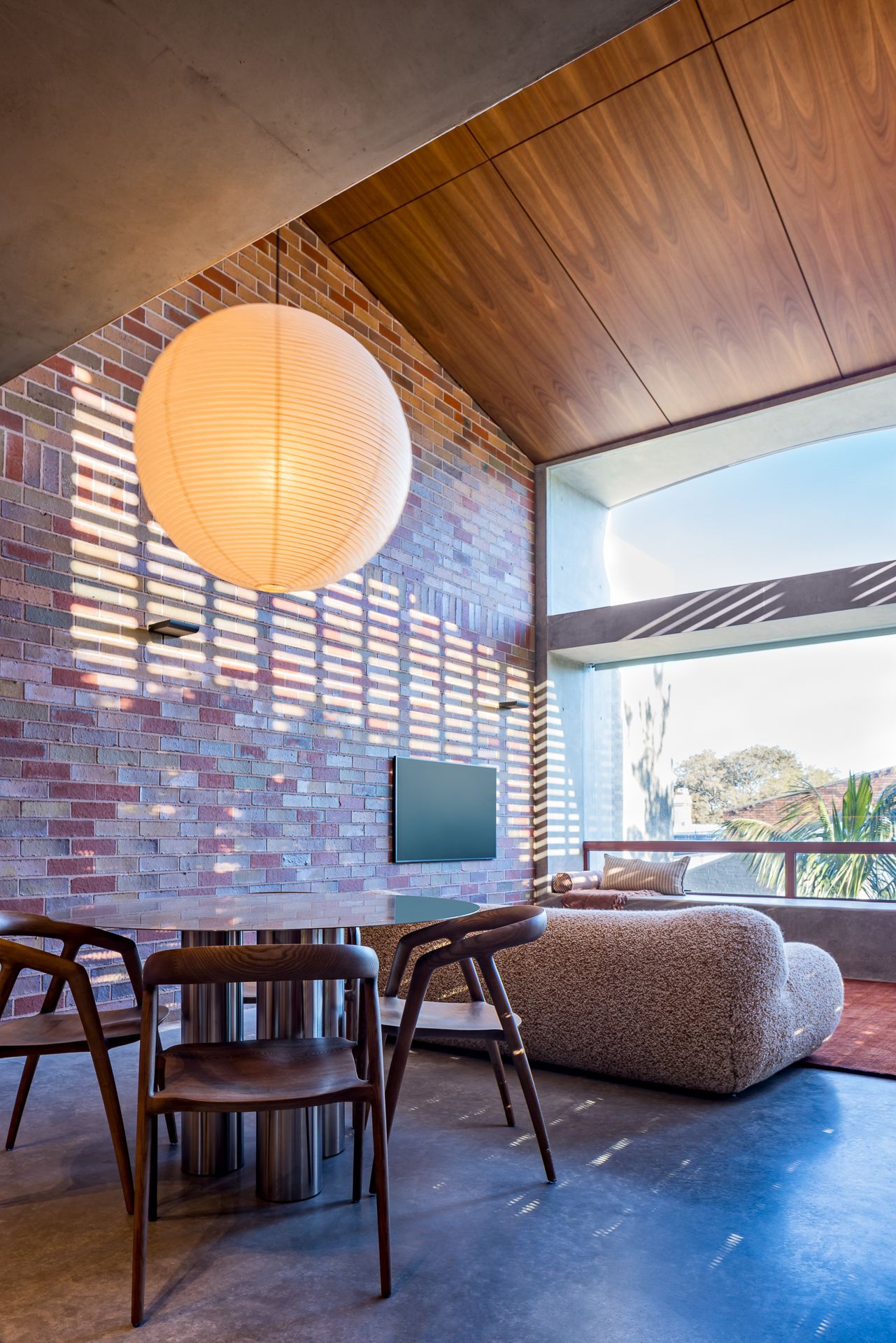 Dining area with round table, timber chairs and large paper lantern beside a brick wall dappled with sunlight through louvres.