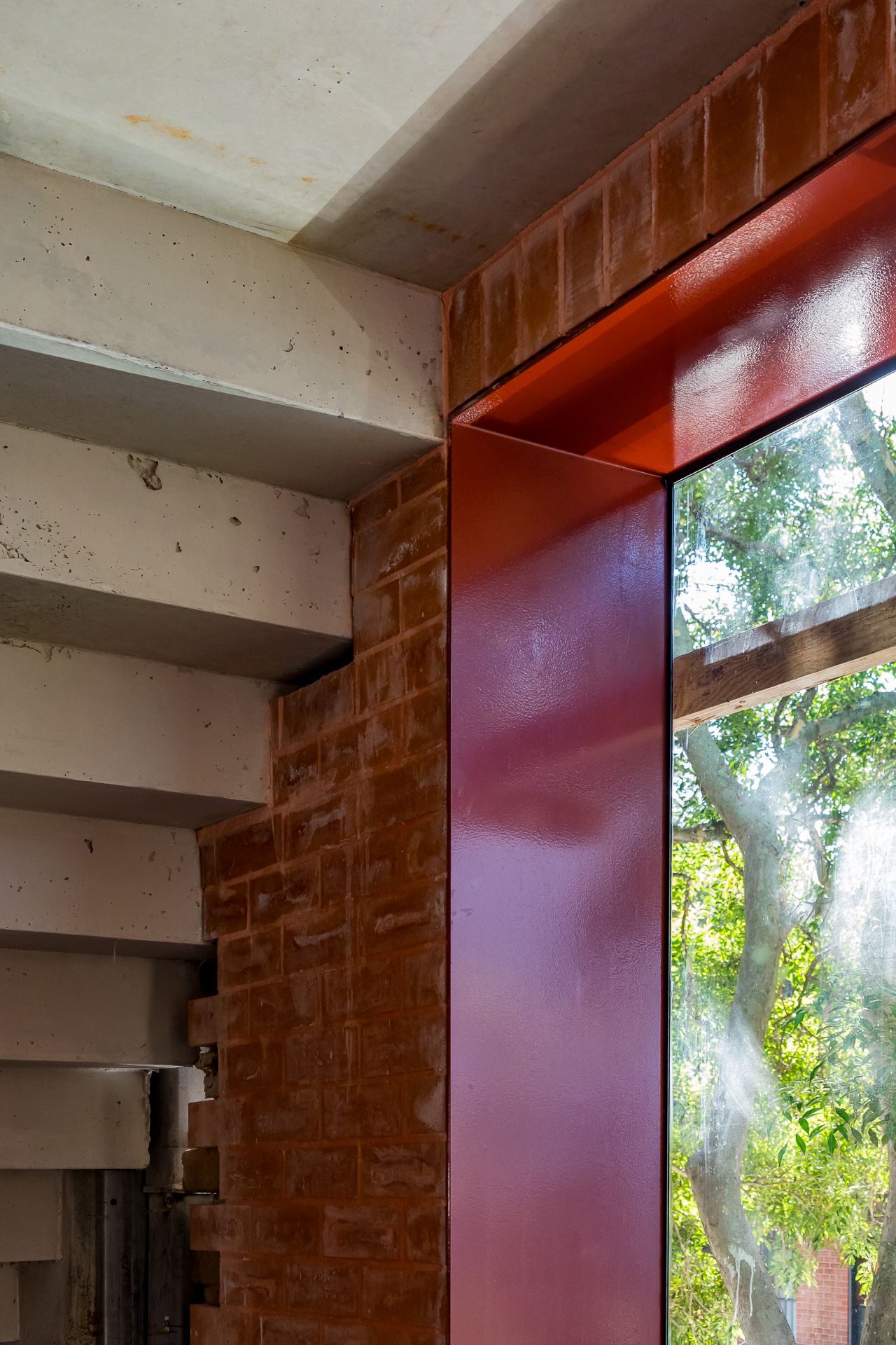 Interior corner with exposed concrete stairs, red brick wall, and a red steel window frame overlooking green trees.