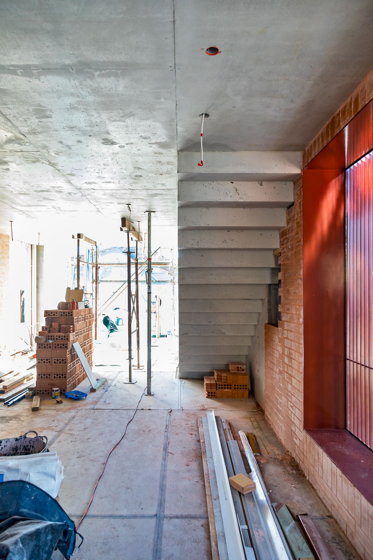 Unfinished building interior under construction with a concrete staircase, stacked bricks, scaffolding and a red-framed window.