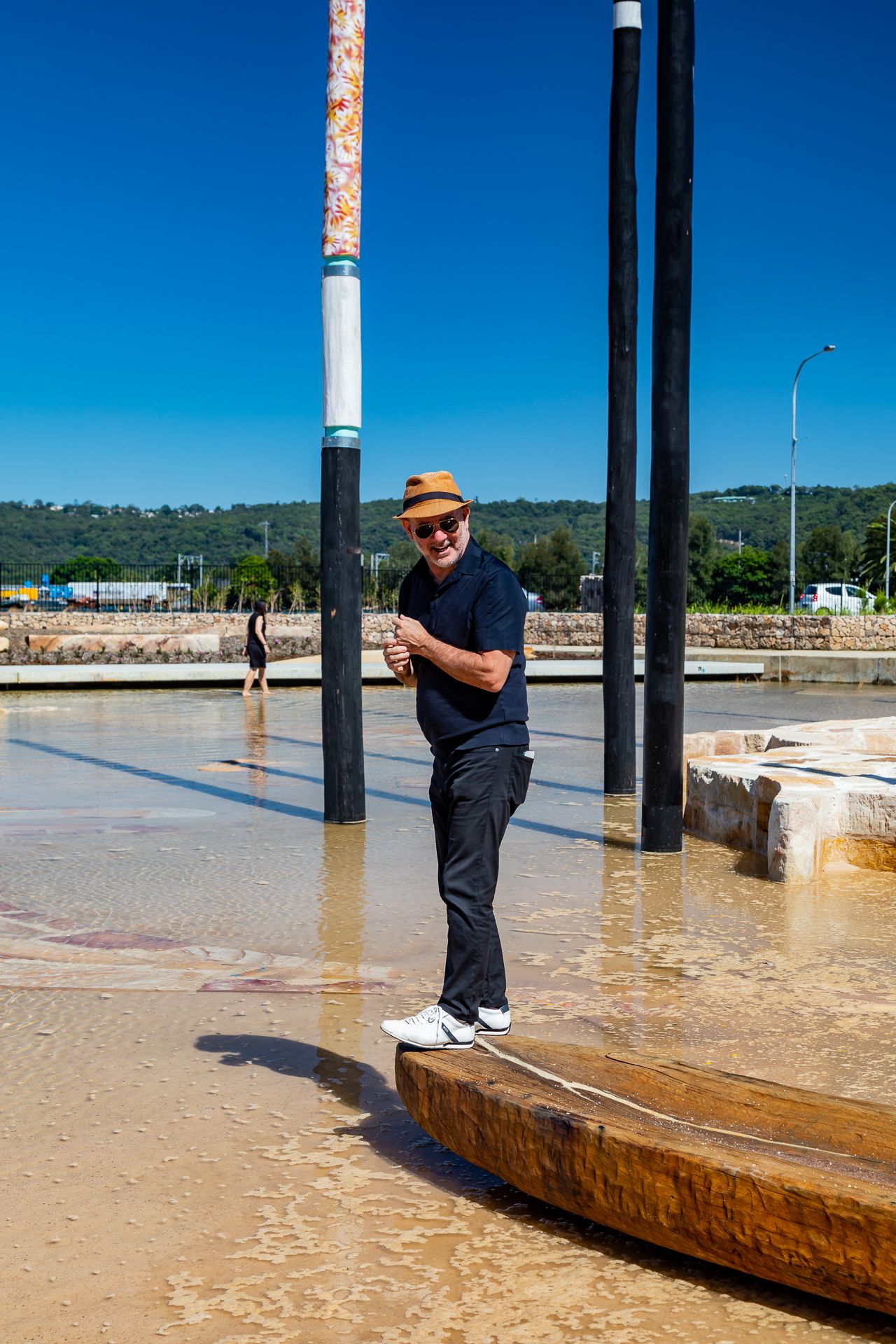 Smiling man in a hat and sunglasses stands in shallow water beside painted poles and a carved timber boat at a waterfront park.