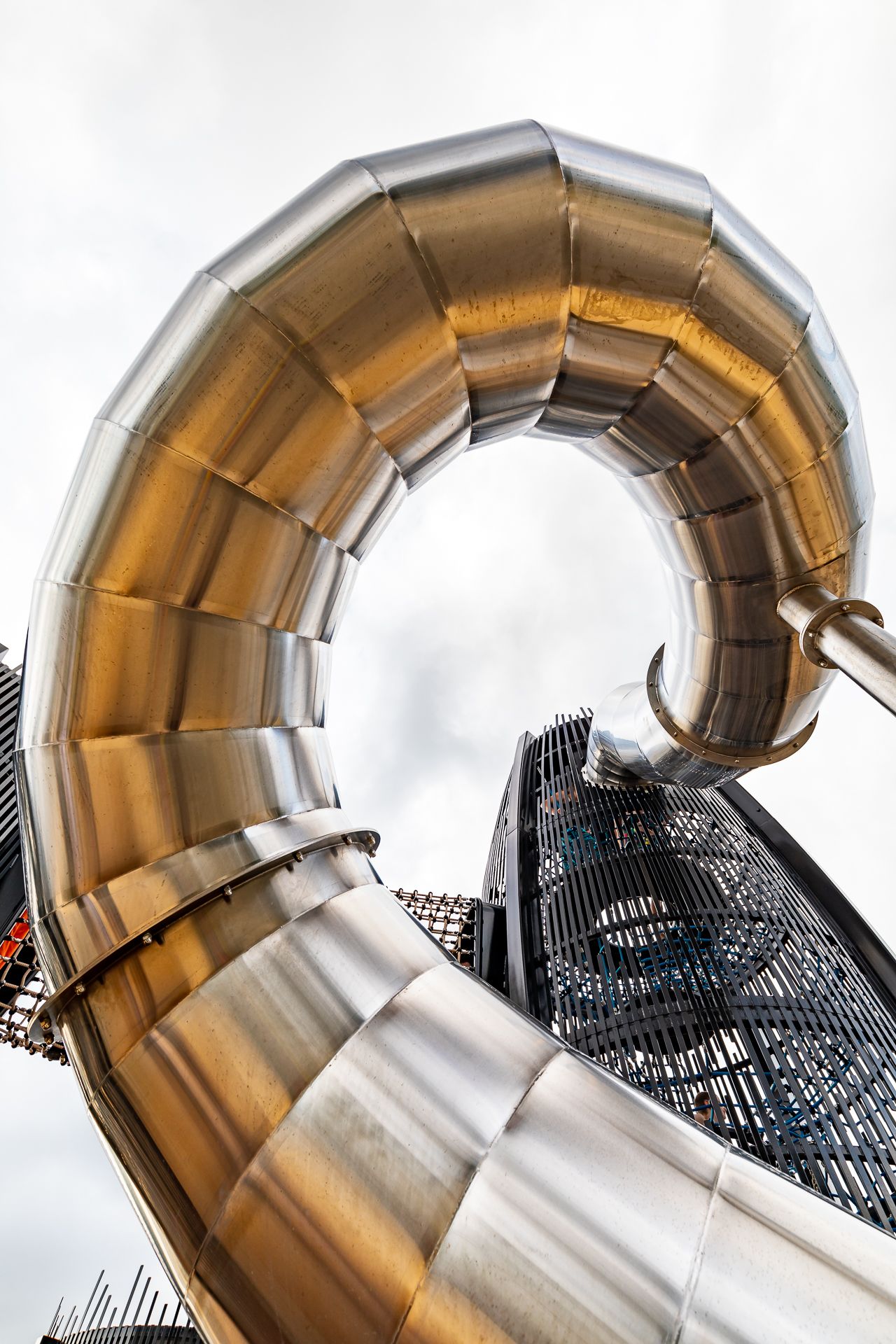 Giant curling stainless steel slide arching over a tall black mesh climbing tower on a bright, cloudy day.