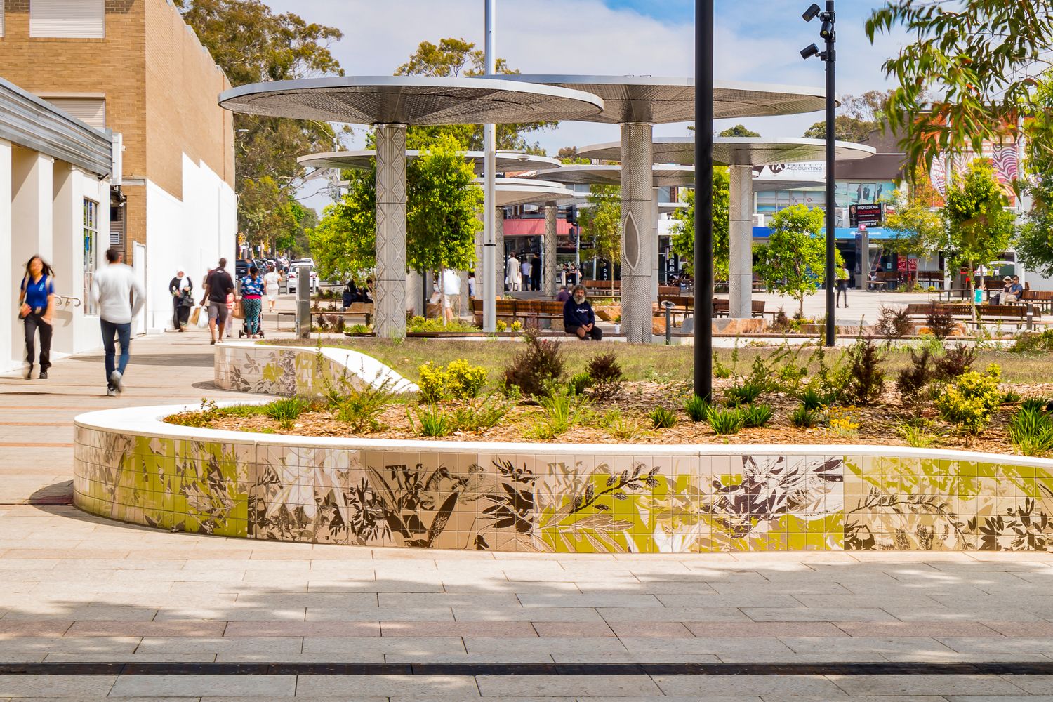 Curved planter wall with painted native motifs beside a shaded public plaza with metal canopies and pedestrians.