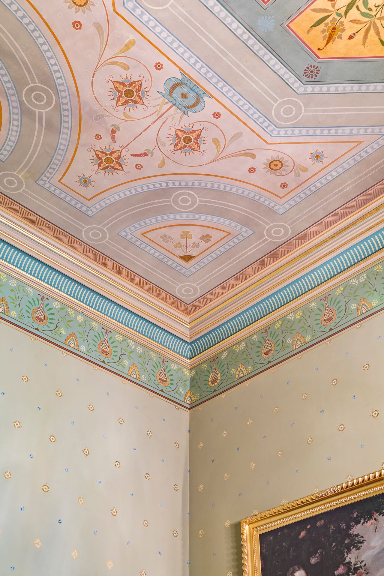 Painted ceiling with floral medallions and decorative frieze above pale walls with gold dots and a gilded picture frame.