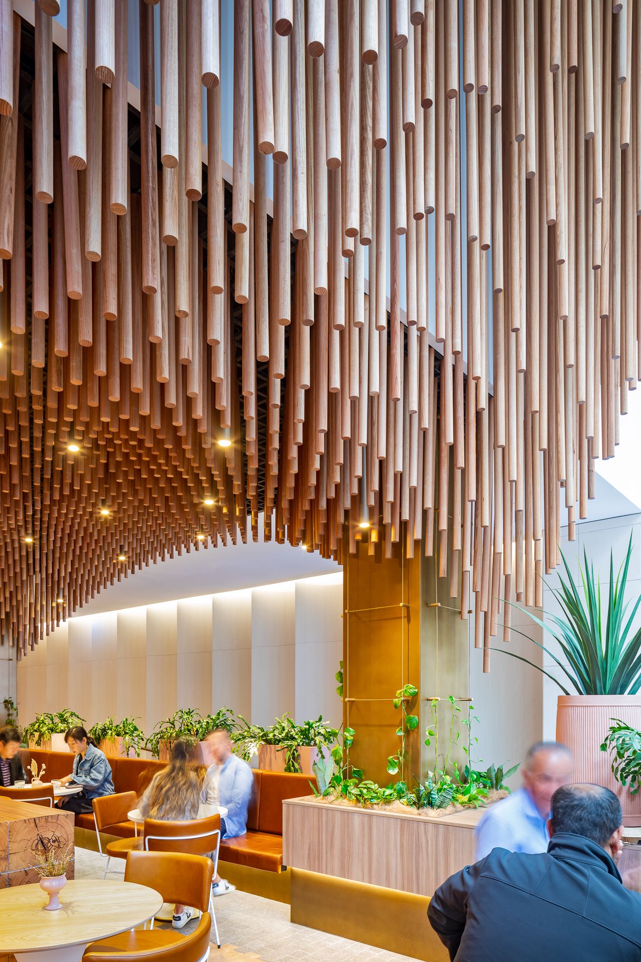Close view of hundreds of suspended timber rods forming a ceiling above diners, tan banquette seating and lush green plants.