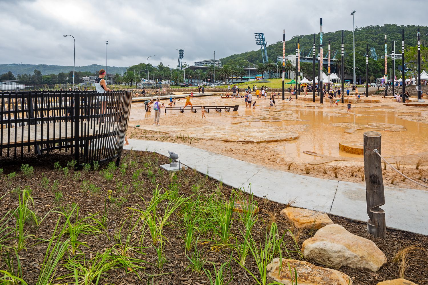A wooden fence and native plantings overlook a shallow water play area with families and event tents in a hillside park.