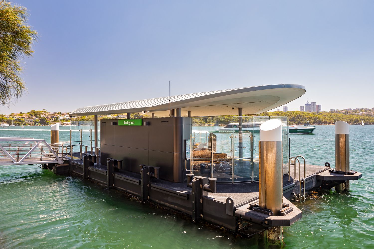 Close view of a modern ferry wharf with a sweeping curved roof, white mooring piles and a gangway over turquoise water beneath a blue sky.
