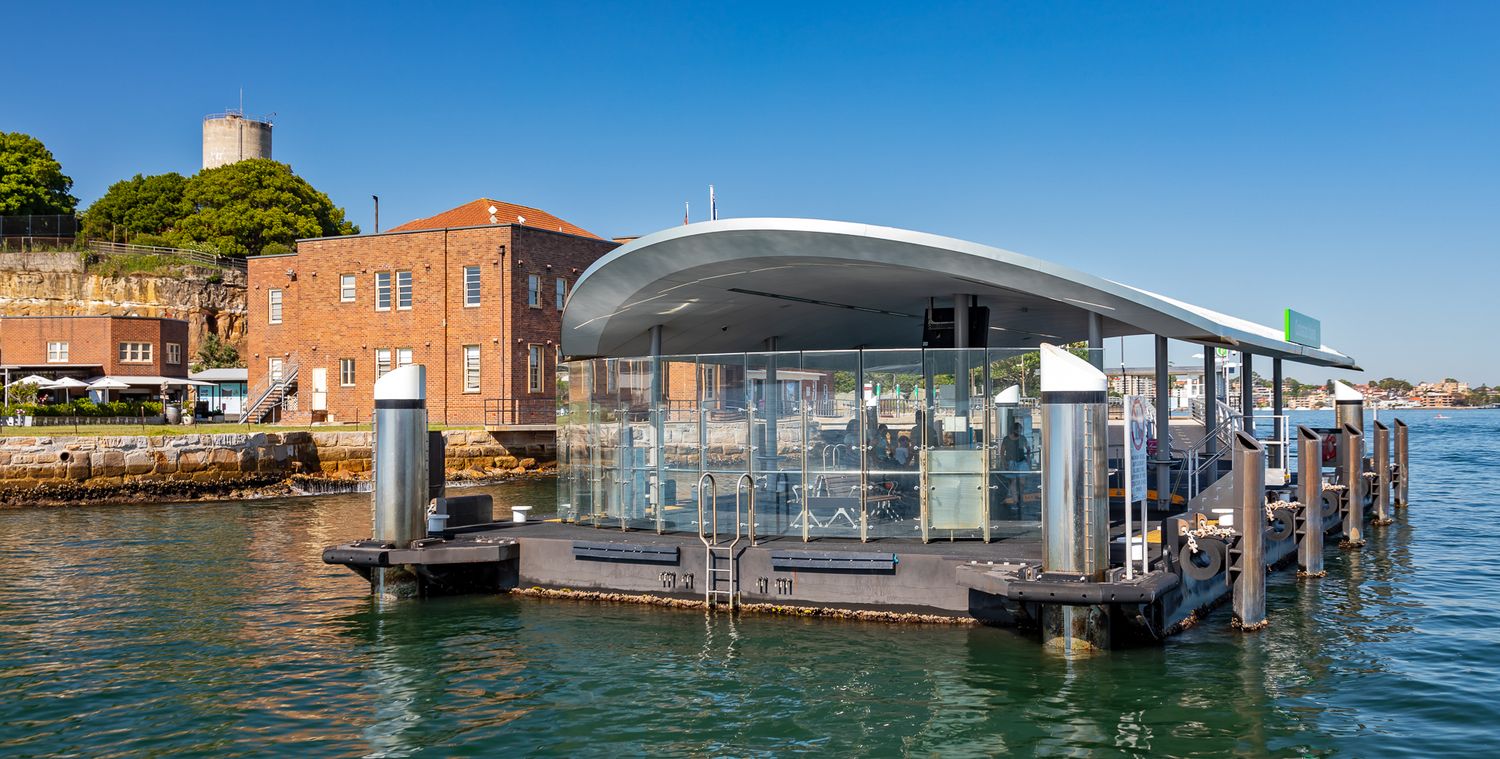 Contemporary ferry terminal with a sweeping curved roof beside a historic brick building and a tower, on sparkling harbour water.
