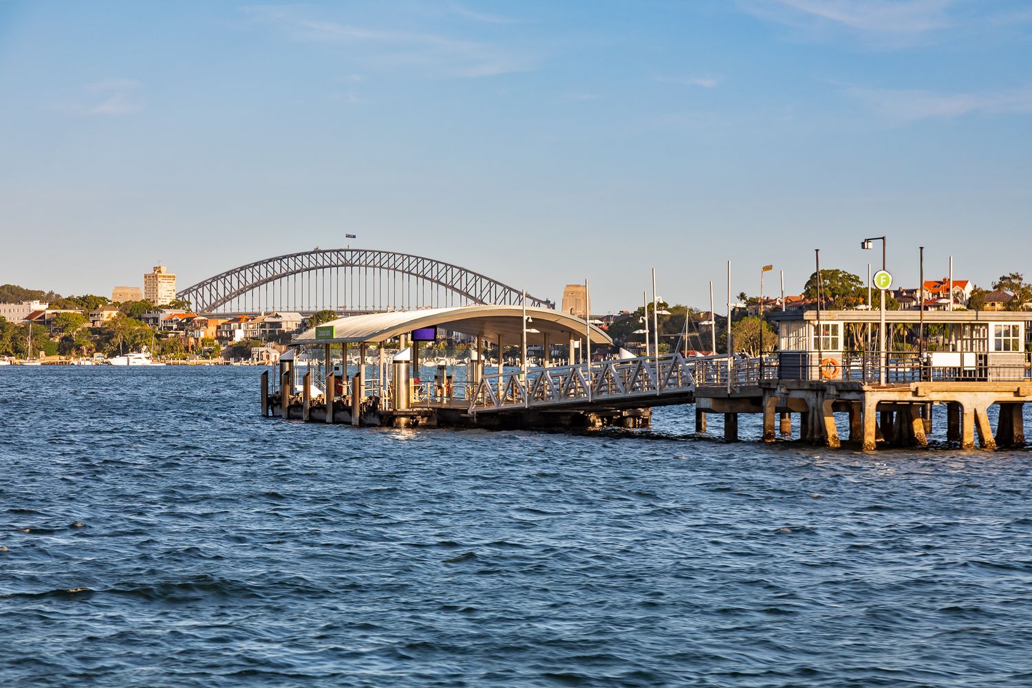 Curved-roof ferry wharf on blue harbour waters, with a steel arch bridge and city skyline visible in the distance.