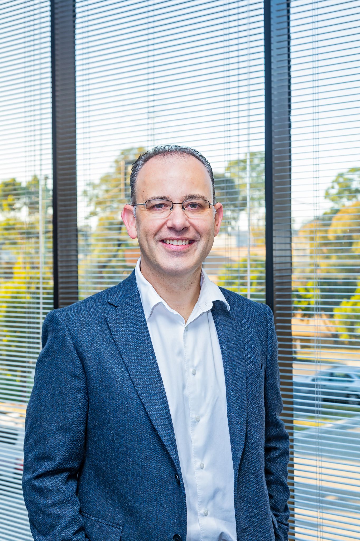 Smiling man with glasses wearing a blue blazer and white open-collared shirt stands beside a large window with blinds and trees outside.