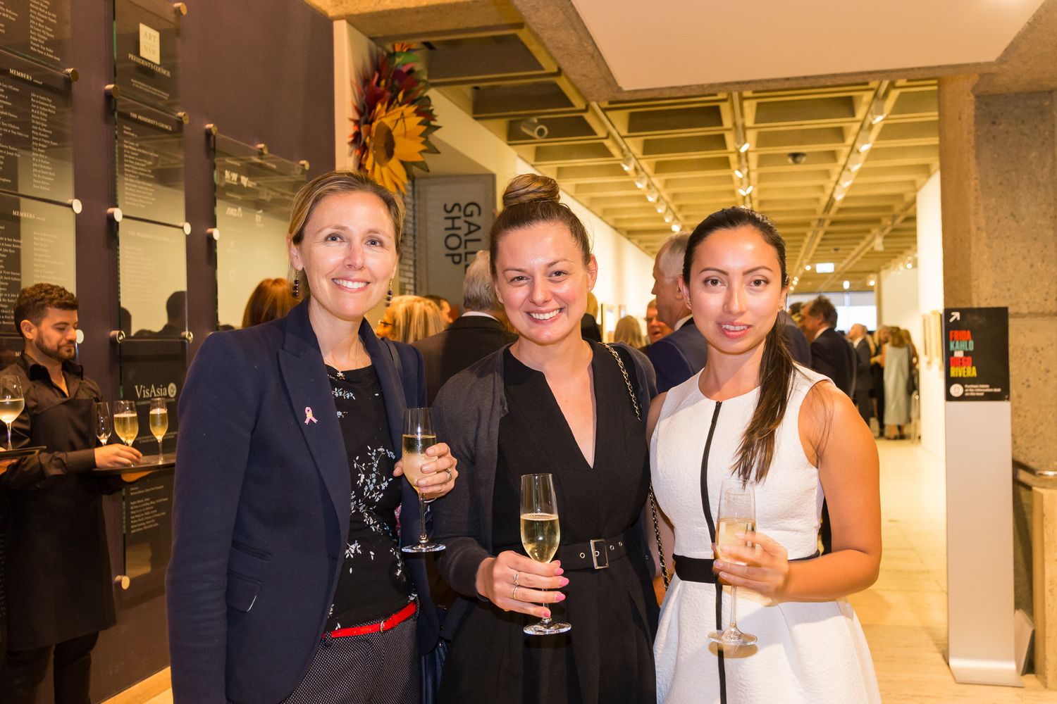Three smiling women hold champagne glasses at a stylish networking event with guests mingling in the background.