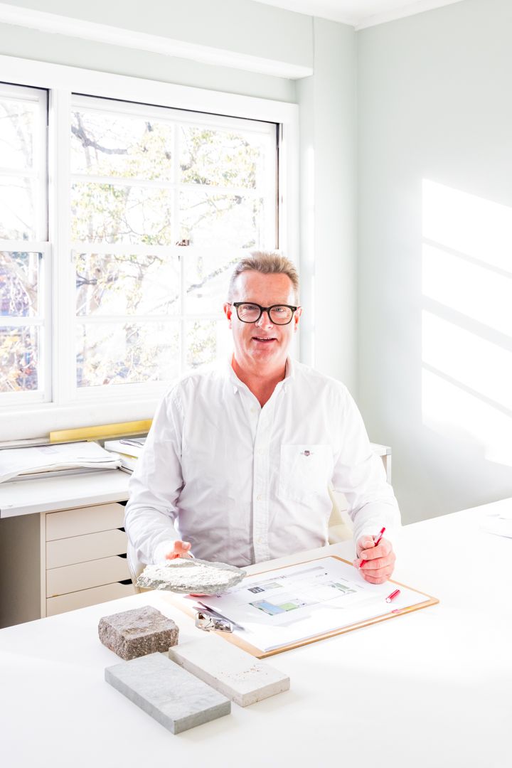 Smiling man in glasses and a white shirt sits at a bright desk reviewing plans with stone and material samples laid out in front of him.