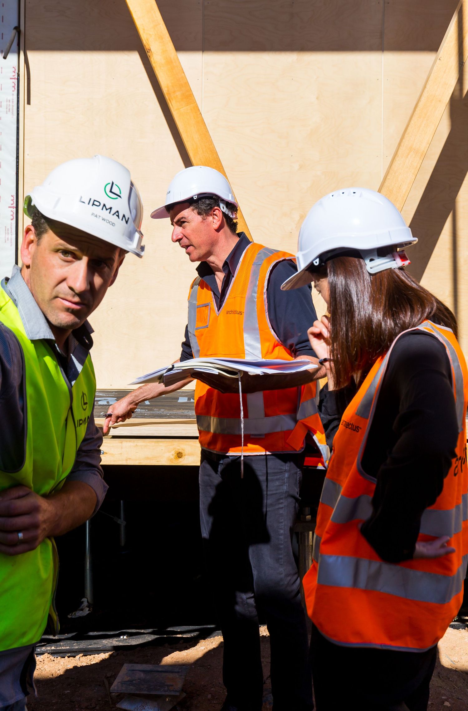Three construction professionals in hard hats and high-visibility vests review plans on site, one wearing a Lipman branded helmet.
