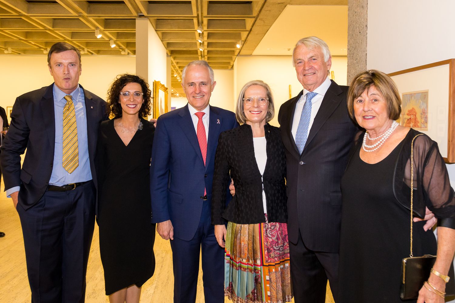Six smiling adults in formal attire pose together for a group photo inside a bright gallery space with framed artwork.