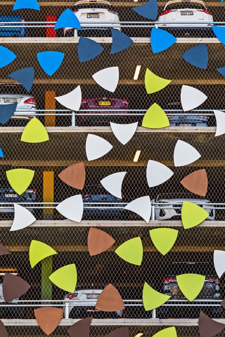 Colorful triangular and rounded shapes in green, blue, white and brown decorate a car park facade with parked cars visible behind mesh.