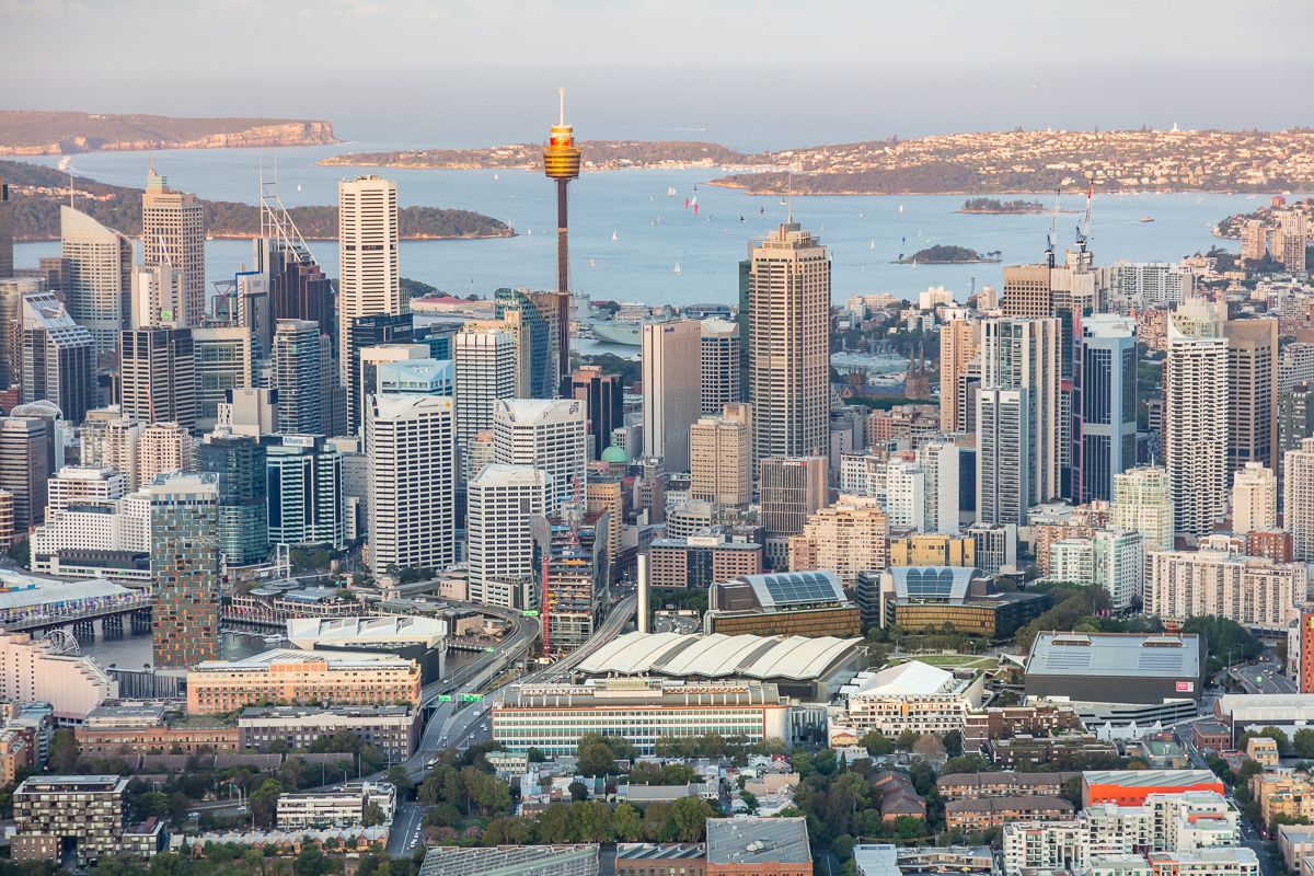 Aerial view of a city skyline with tall office towers, a prominent observation tower and a harbour in the background at dusk.