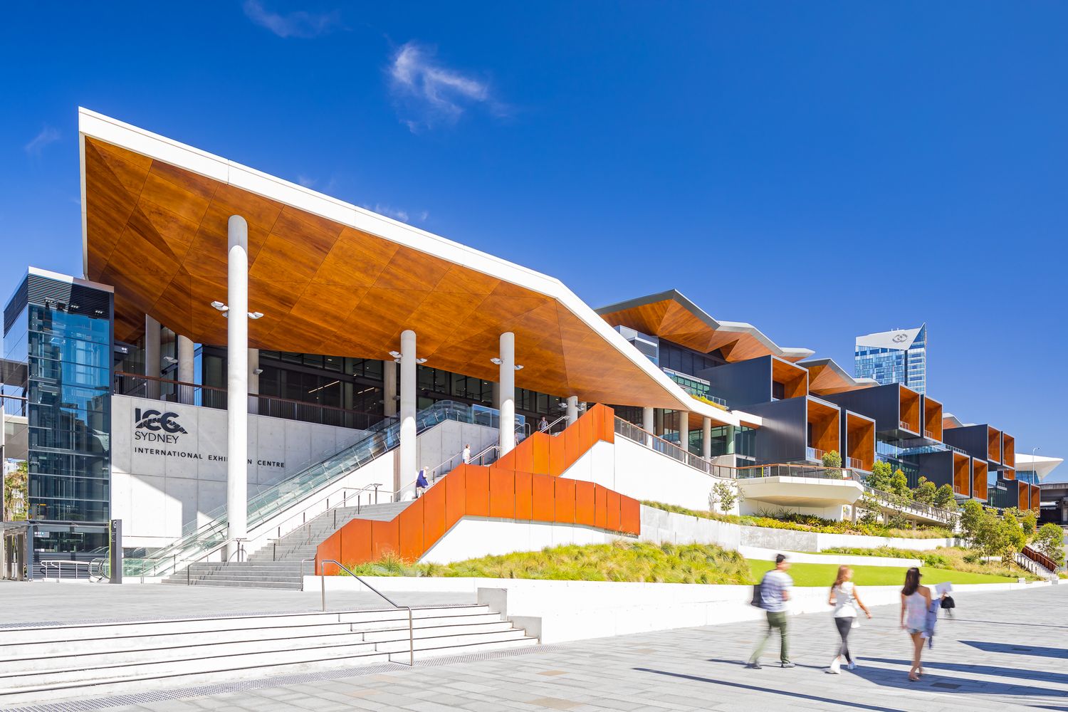 Pedestrians walk past a convention centre entrance with a timber-lined roof canopy, white columns, an orange ramp and wide stone steps.