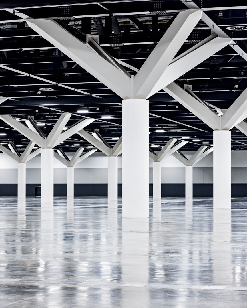 White branching structural columns rise from a glossy reflective floor in a spacious modern exhibition hall.