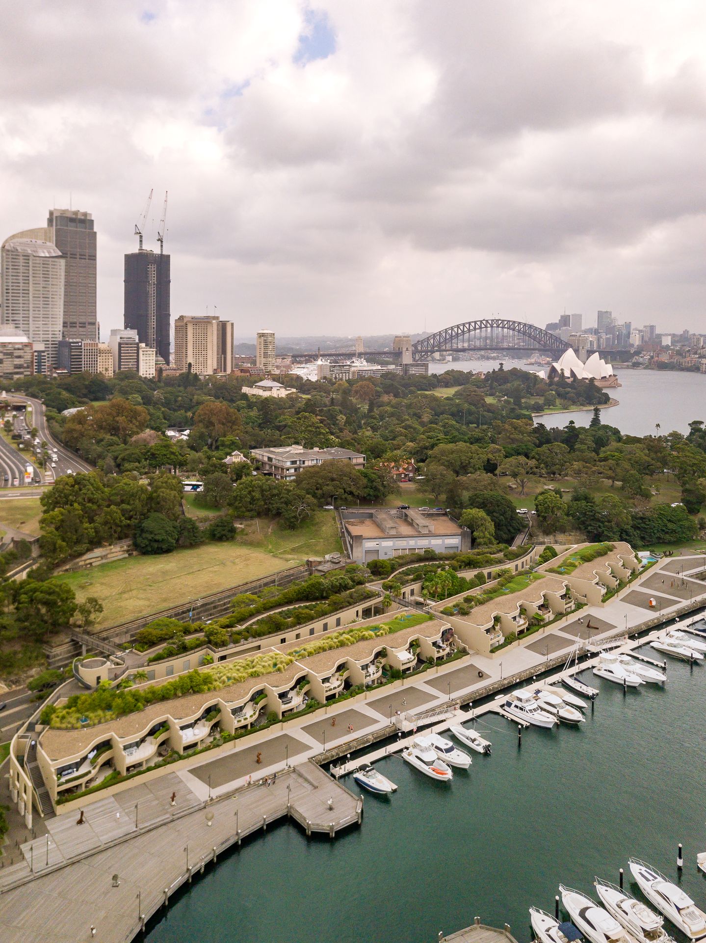 Aerial view of a harbourside waterfront with terraced buildings, moored boats, city skyline and the Sydney Harbour Bridge beyond.