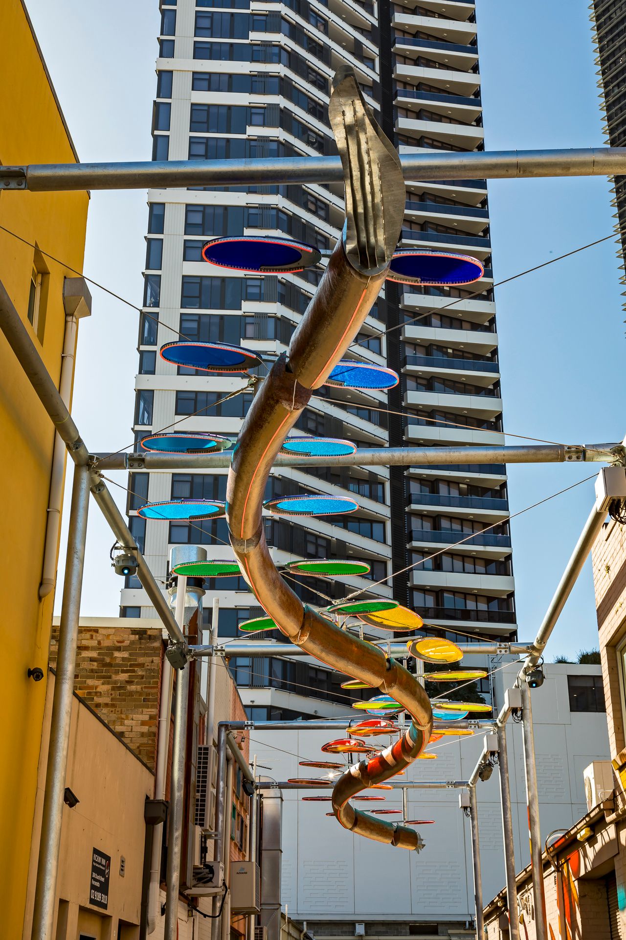 Spiraling public art installation with colorful translucent discs hanging over a city alley, framed by a high-rise tower.