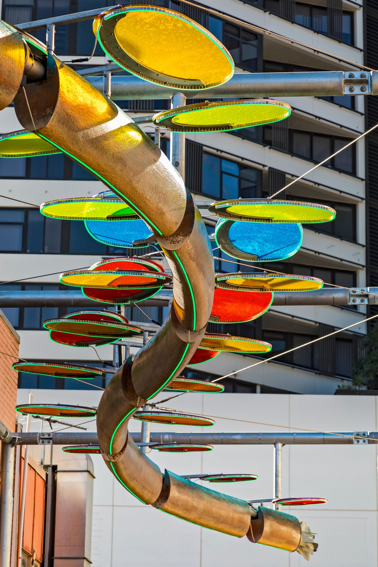 Colorful translucent discs in yellow, green, blue and red surround a curving metallic tube sculpture suspended above an urban street.