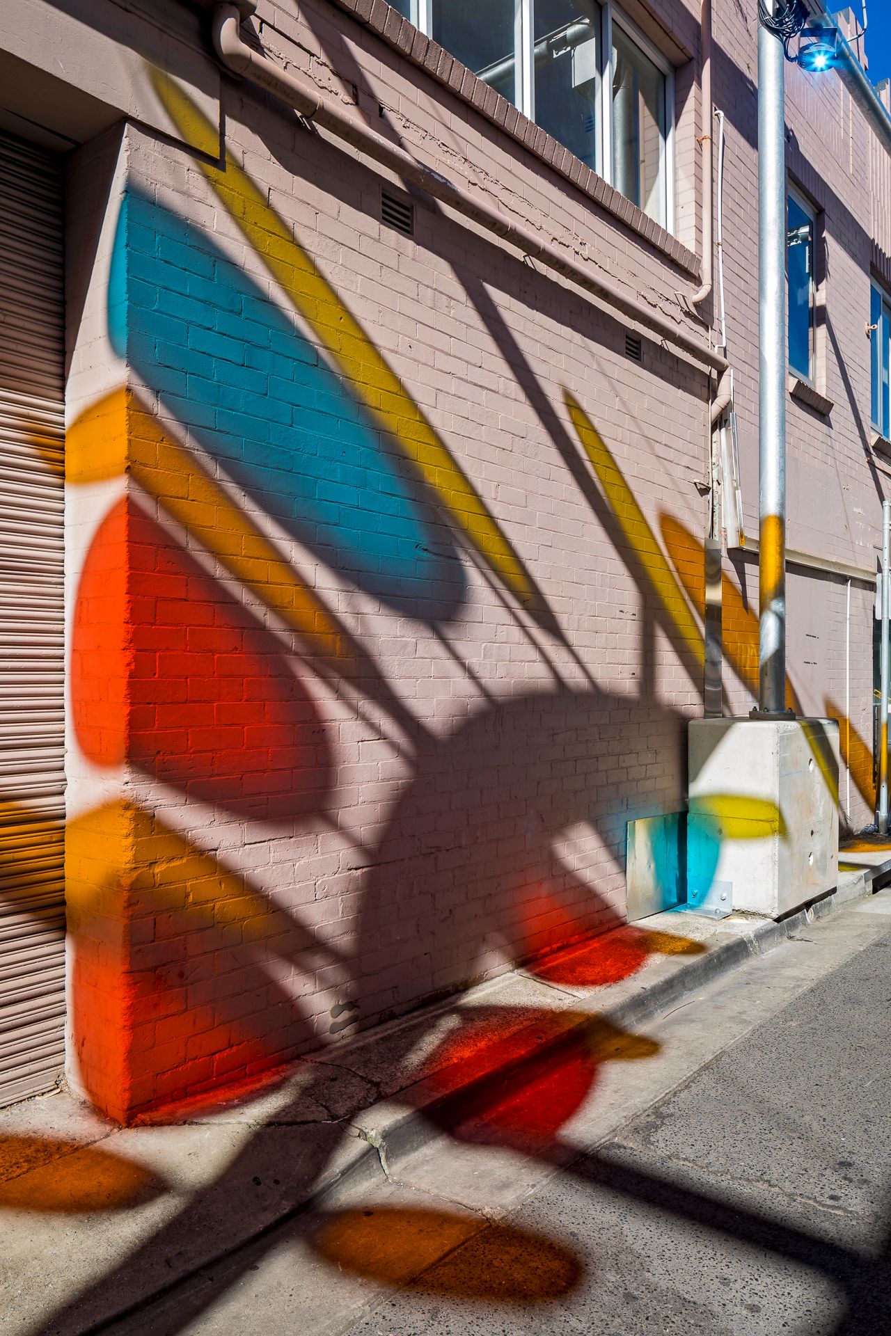 Close view of a brick building corner with bold blue, orange and yellow geometric shapes and dramatic shadows on the wall.