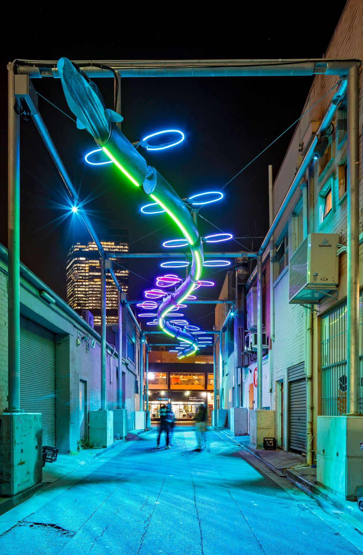 Blurred figures move through a brick laneway at night under a spiral light sculpture glowing in blue, green and purple neon rings.