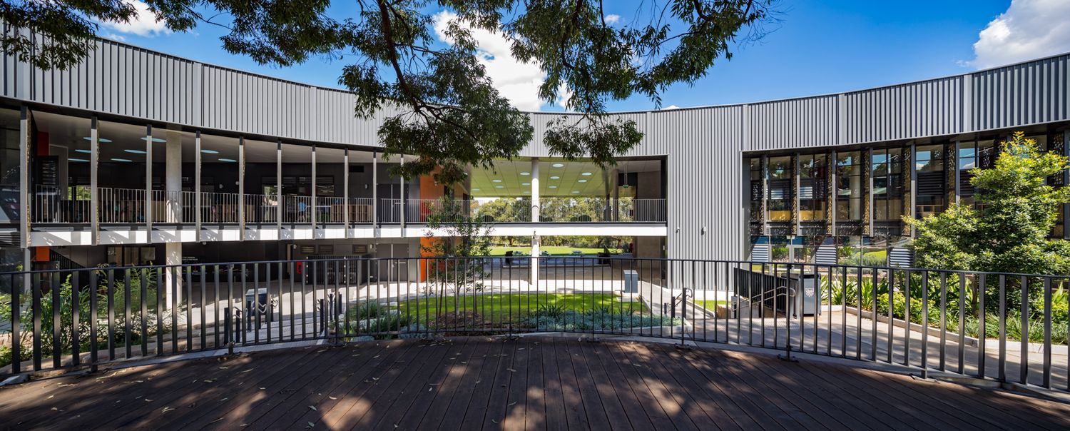 Modern two-storey building with a clerestory-lit courtyard, timber-framed glazing and leafy trees viewed through a metal balustrade.