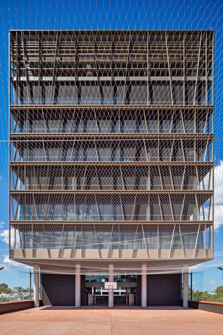 Multi-storey building with a layered metal mesh facade and exposed structure rising above a terracotta courtyard against a blue sky.