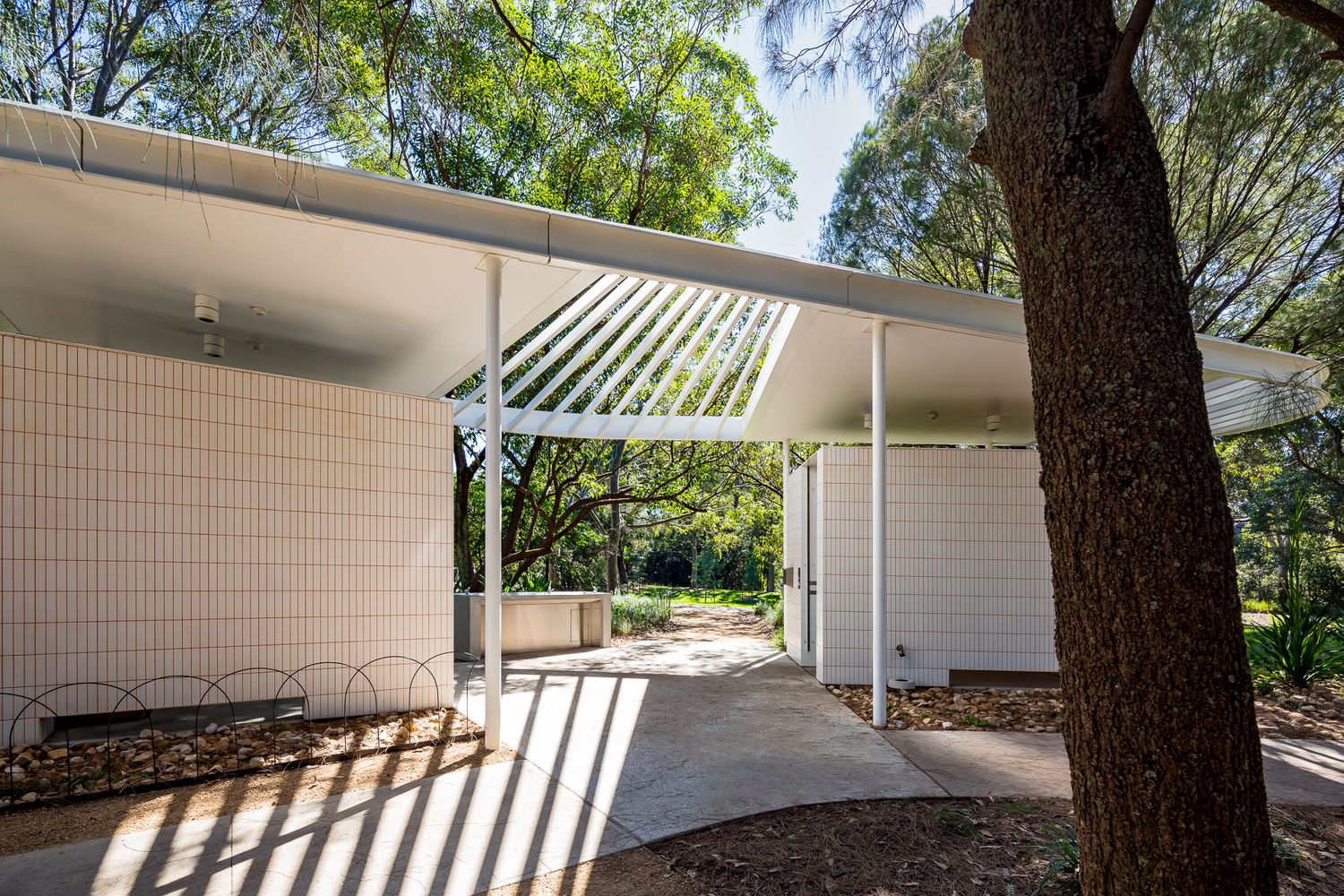 Sunlit walkway beside a white tiled restroom block with a sleek overhanging roof and slender poles, framed by trees.