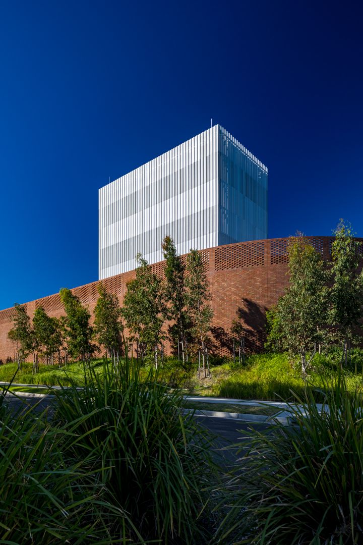 Modern white louvered tower rising behind a curved brick wall with young trees and tall grasses under a deep blue sky.