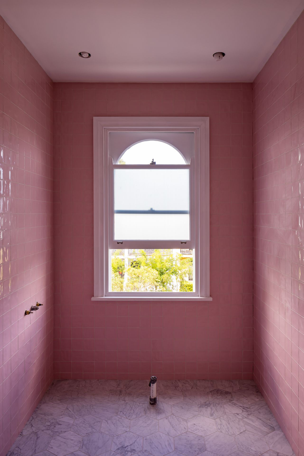 Pink-tiled bathroom with a white sash window with arched top, marble hexagon floor tiles and recessed ceiling lights.