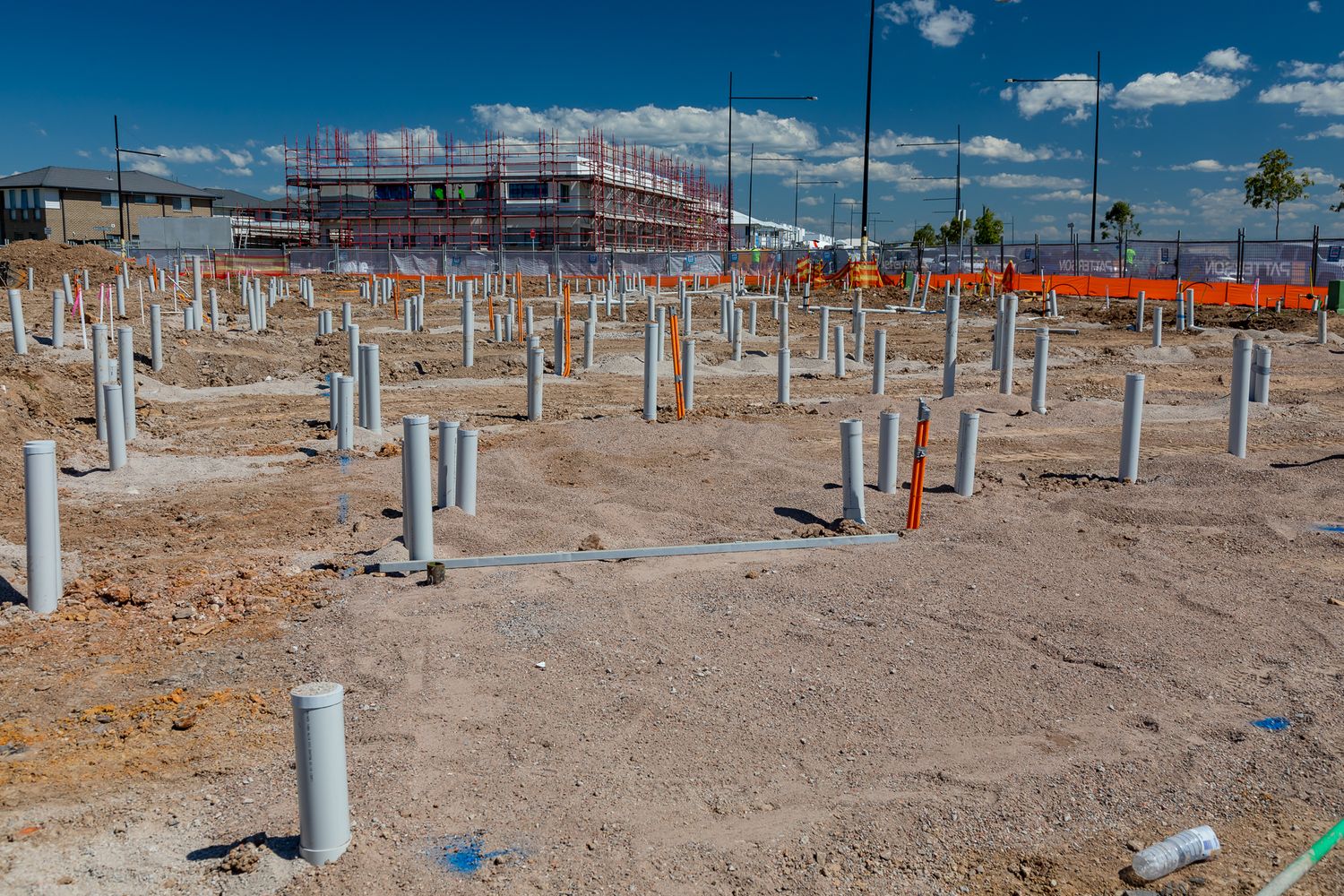 Rows of white plumbing pipes protruding from bare ground at a residential building site with a multi-storey frame in the background.