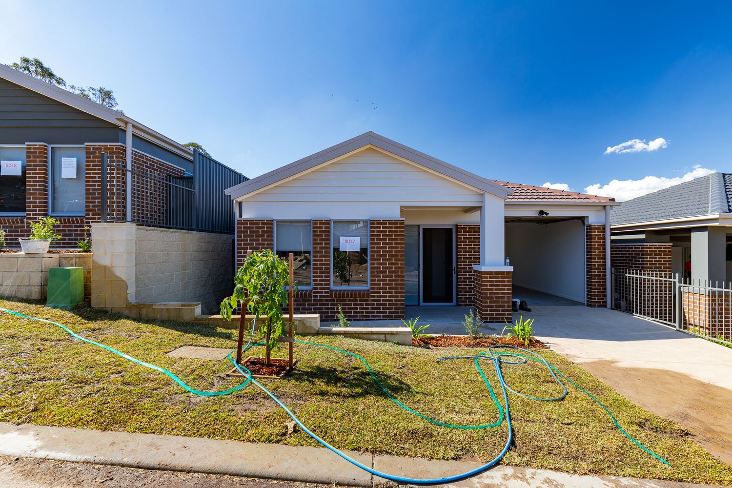 Newly built brick single-storey home with a carport, young sapling, fresh turf and a green hose in the front yard.