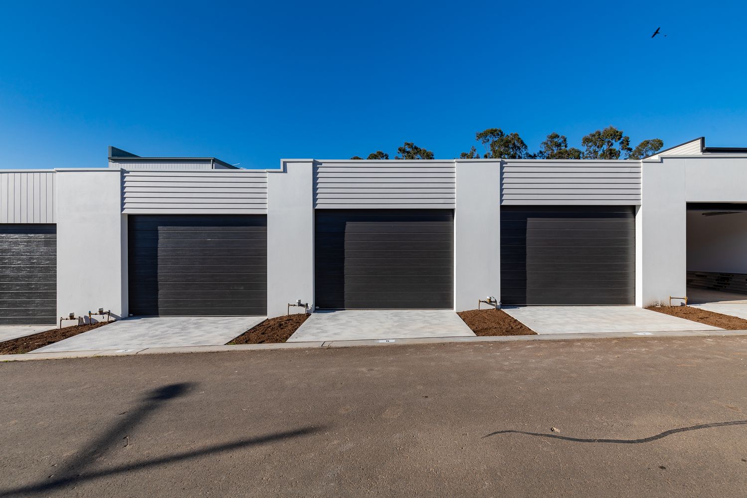 Row of modern townhouses with dark roller garage doors, concrete driveways and light gray cladding under a bright blue sky.