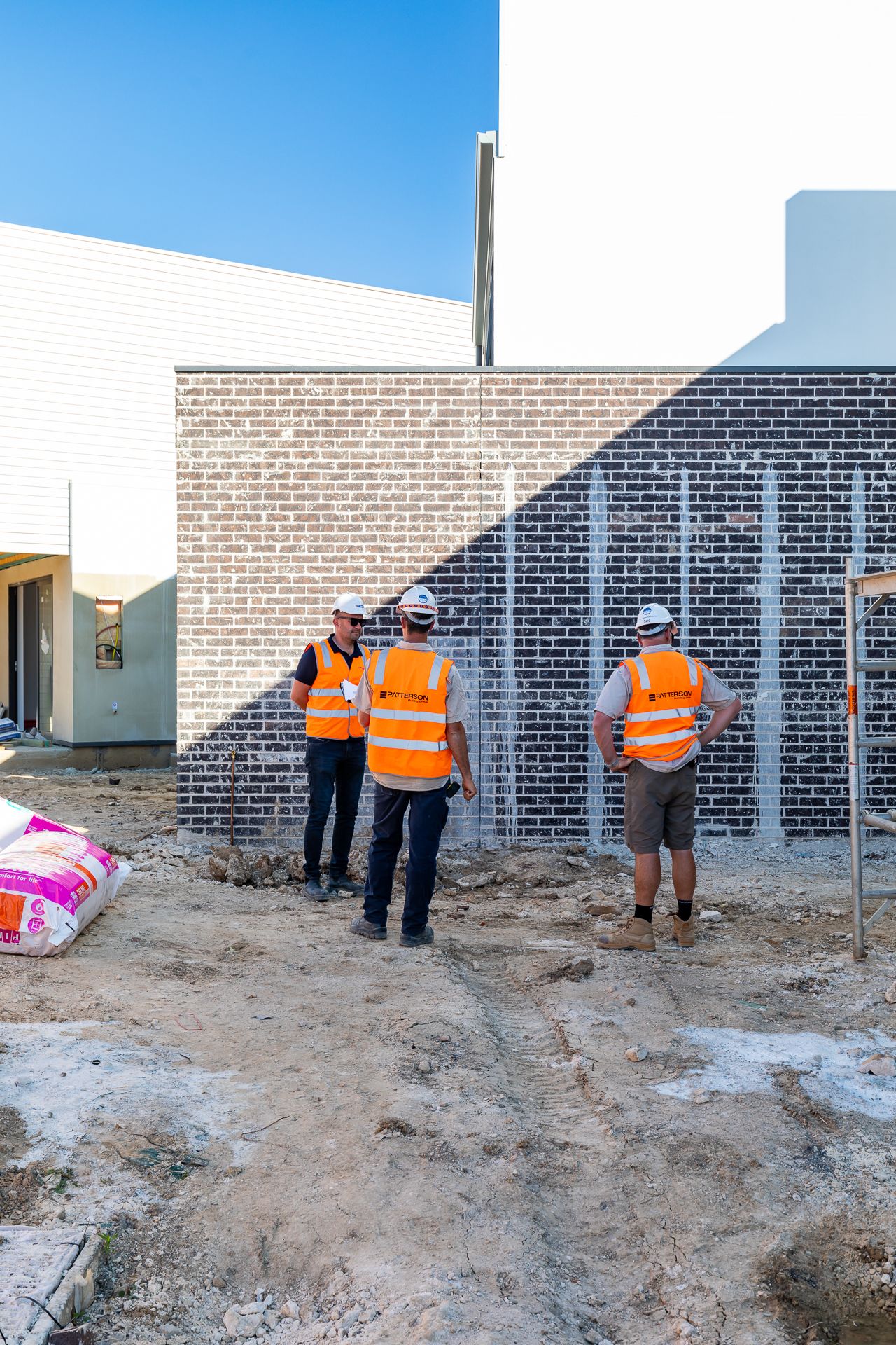 Three construction workers in orange hi-vis vests and hard hats inspect a dark brick wall at a building site.