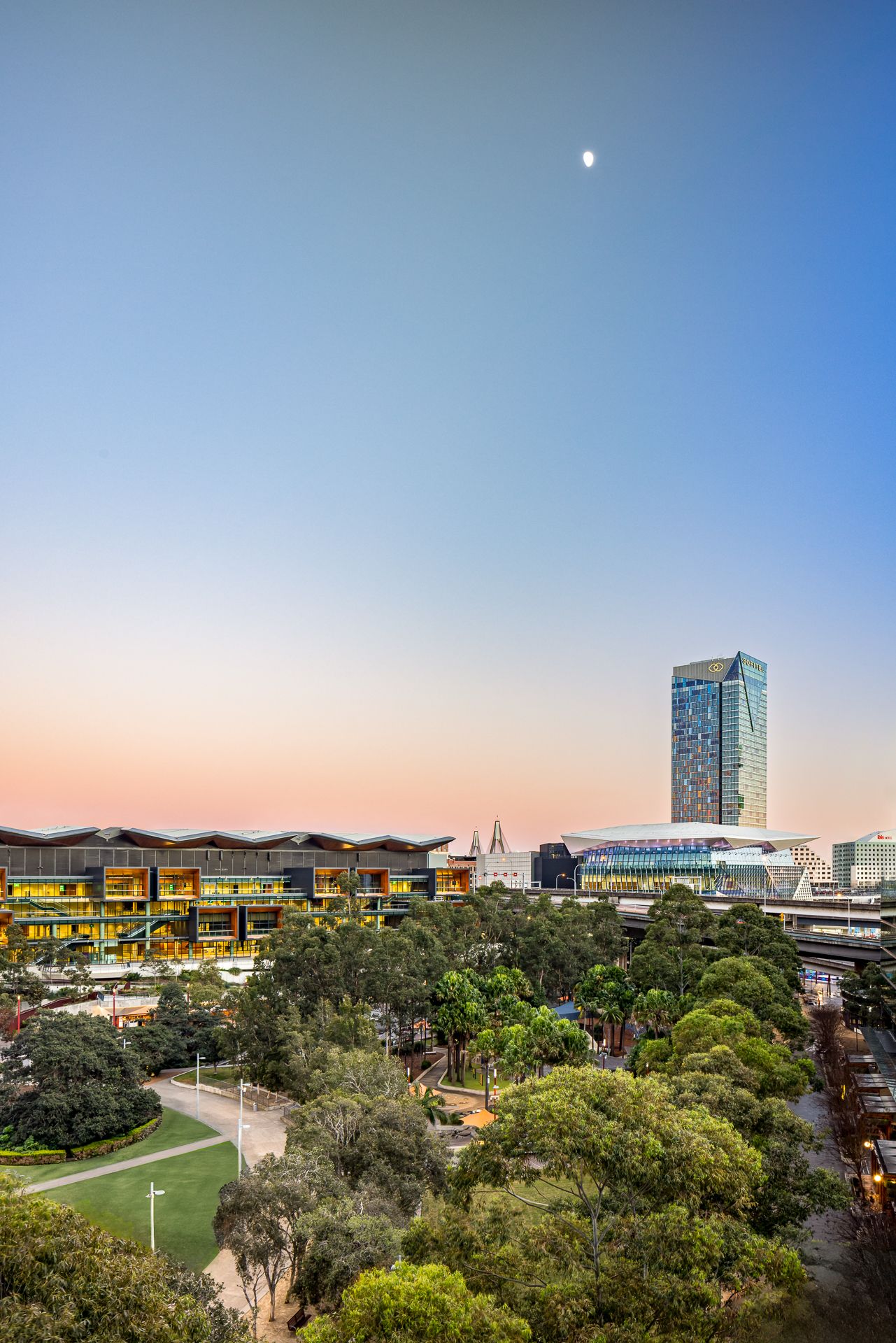 Twilight view over parkland trees toward a modern convention centre and glass skyscraper under a pink sky with a rising moon.