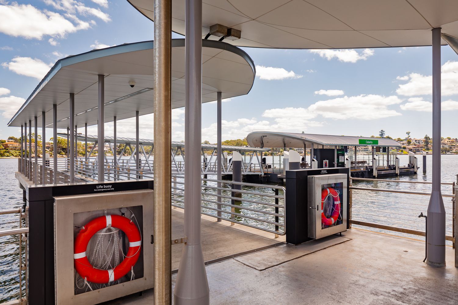 Ferry wharf platform with lifebuoy cabinets, curved roof canopies and railings overlooking the harbour on a sunny day.