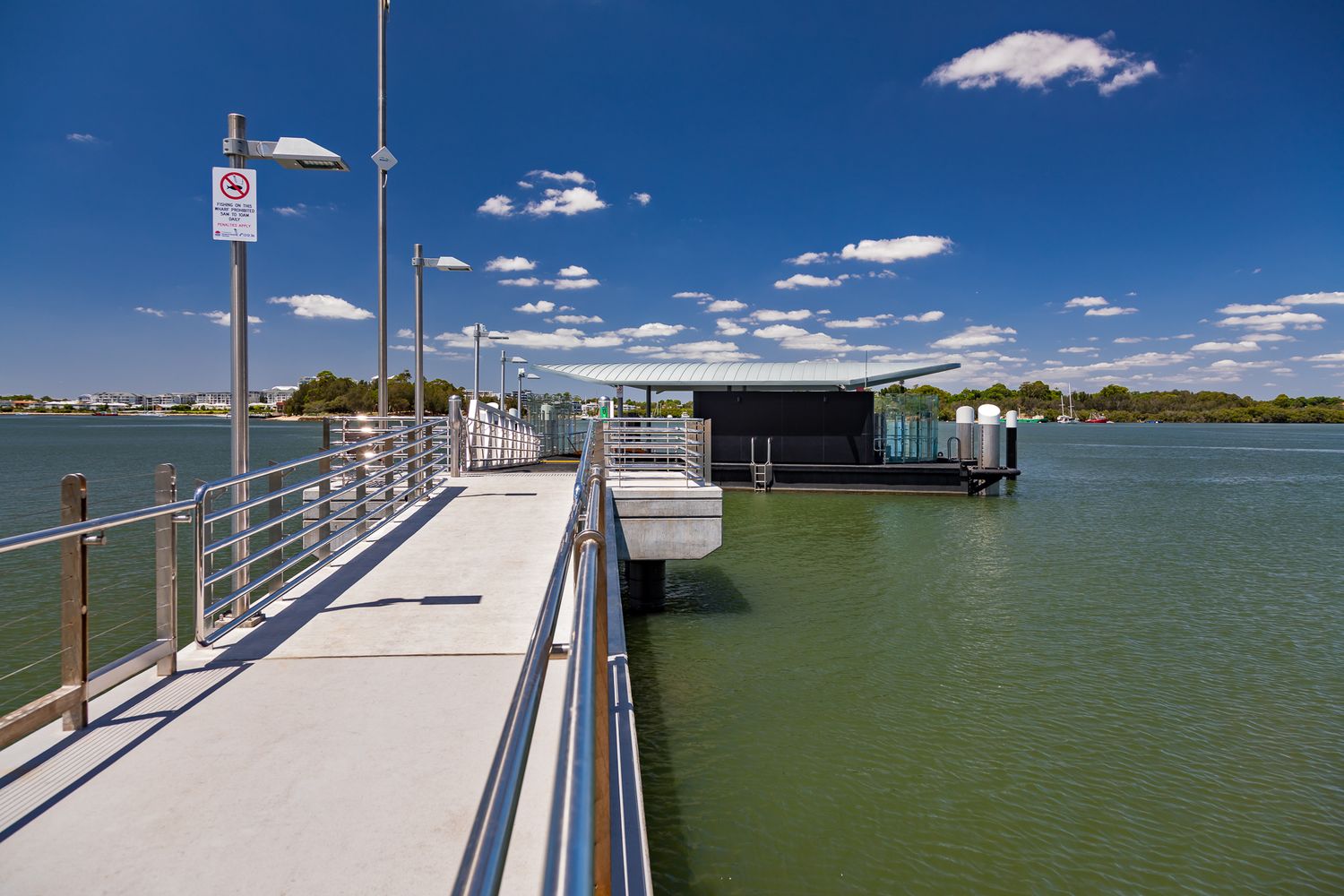 Concrete walkway with metal railings leads to the Kissing Point ferry wharf over green river water under a blue sky.