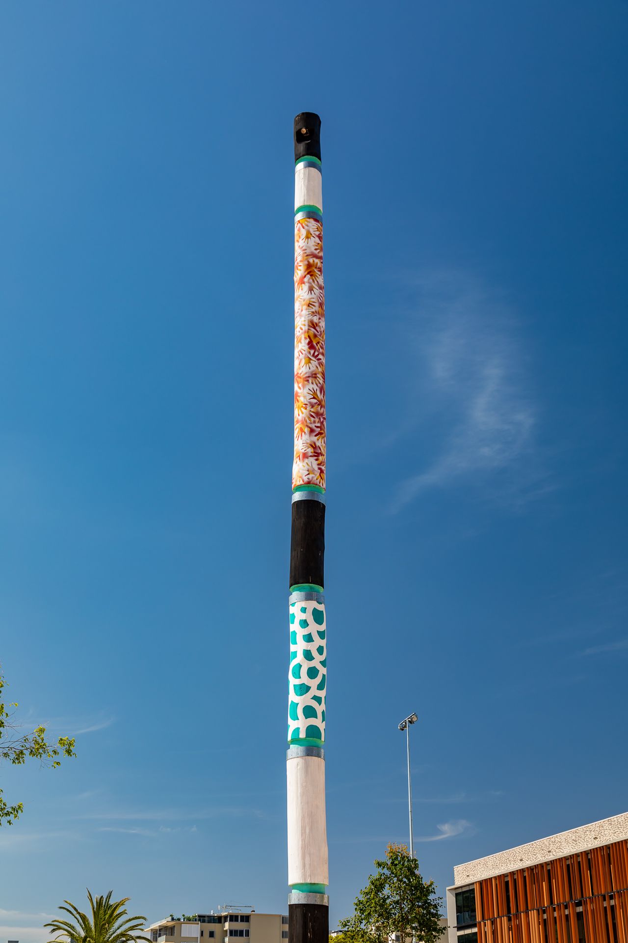 Tall slender painted pole with Aboriginal-style patterns in white, black, teal and orange rising against a bright blue sky.