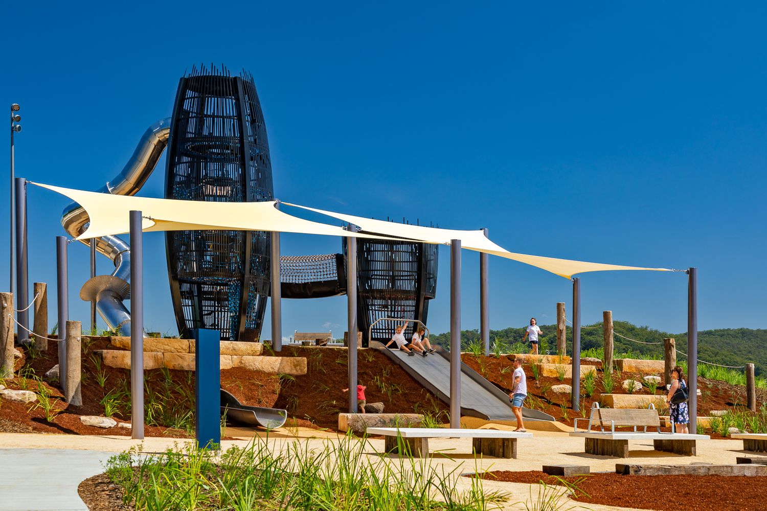 Children play beneath white shade sails at a waterfront playground with a tall black lattice tower and metal slide.