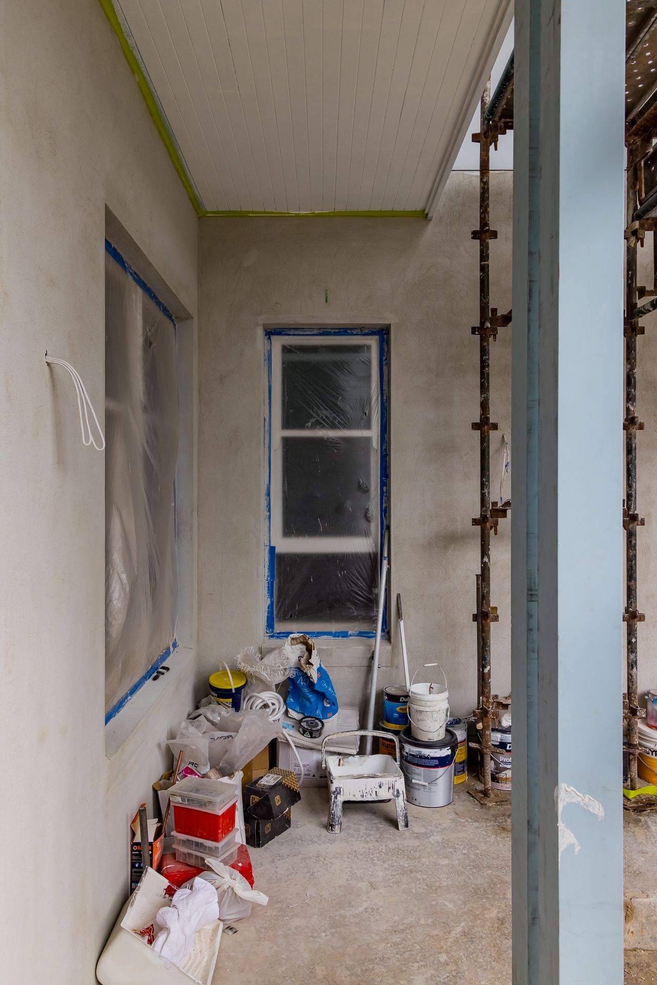 Unfinished room mid-renovation with a taped window, paint buckets, a step ladder, supplies and scaffolding visible beside a blue column.