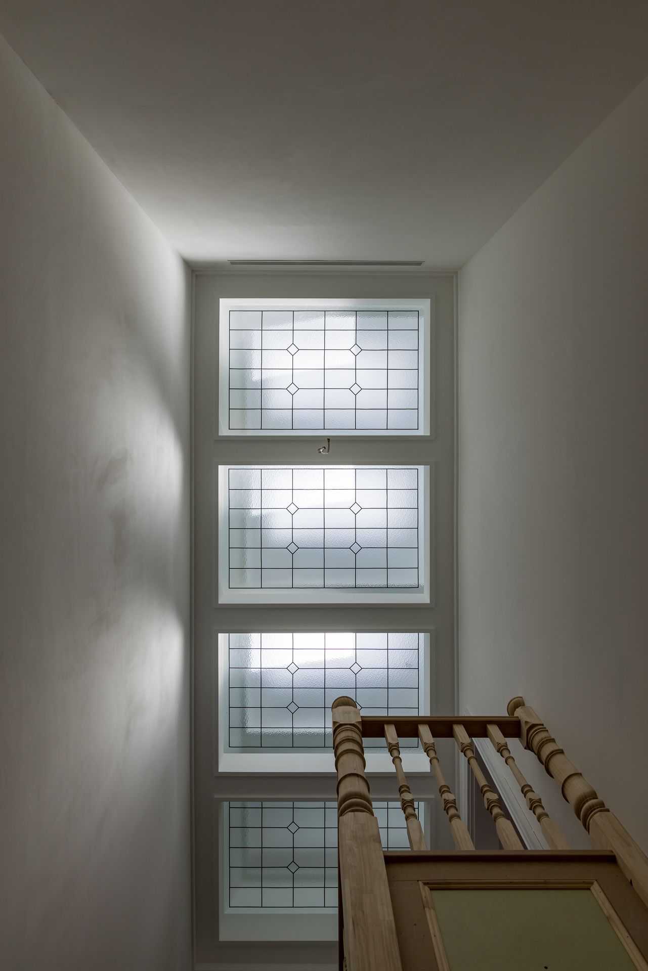Tall stairwell window with leadlight stained glass panels above a timber balustrade, casting soft natural light onto white walls.