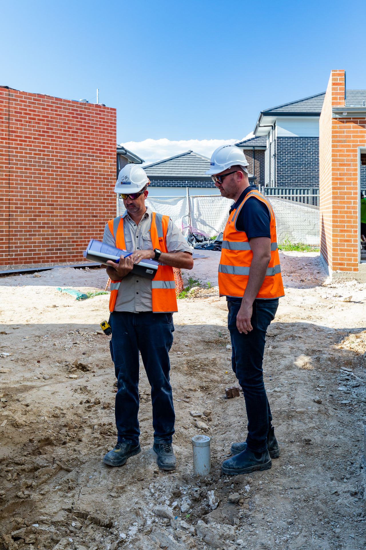 Two construction professionals in hard hats and orange safety vests review plans on a residential building site near new brick walls.