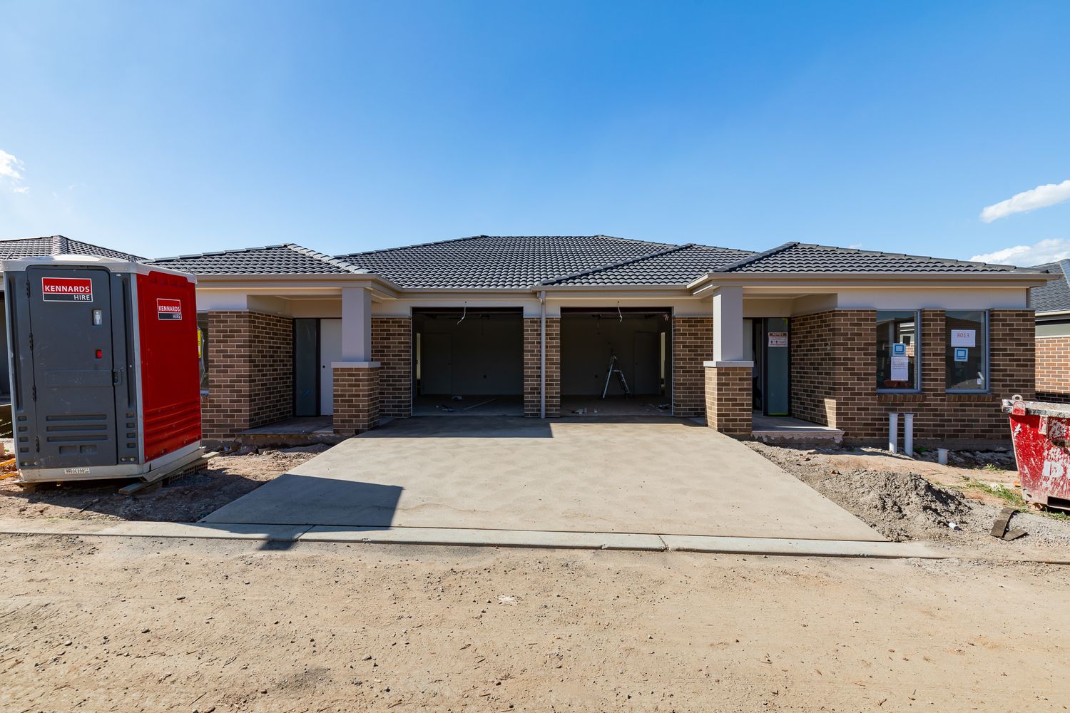 Newly built brick duplex homes with tiled roofs and open garages on a dirt building lot with a portable toilet nearby.