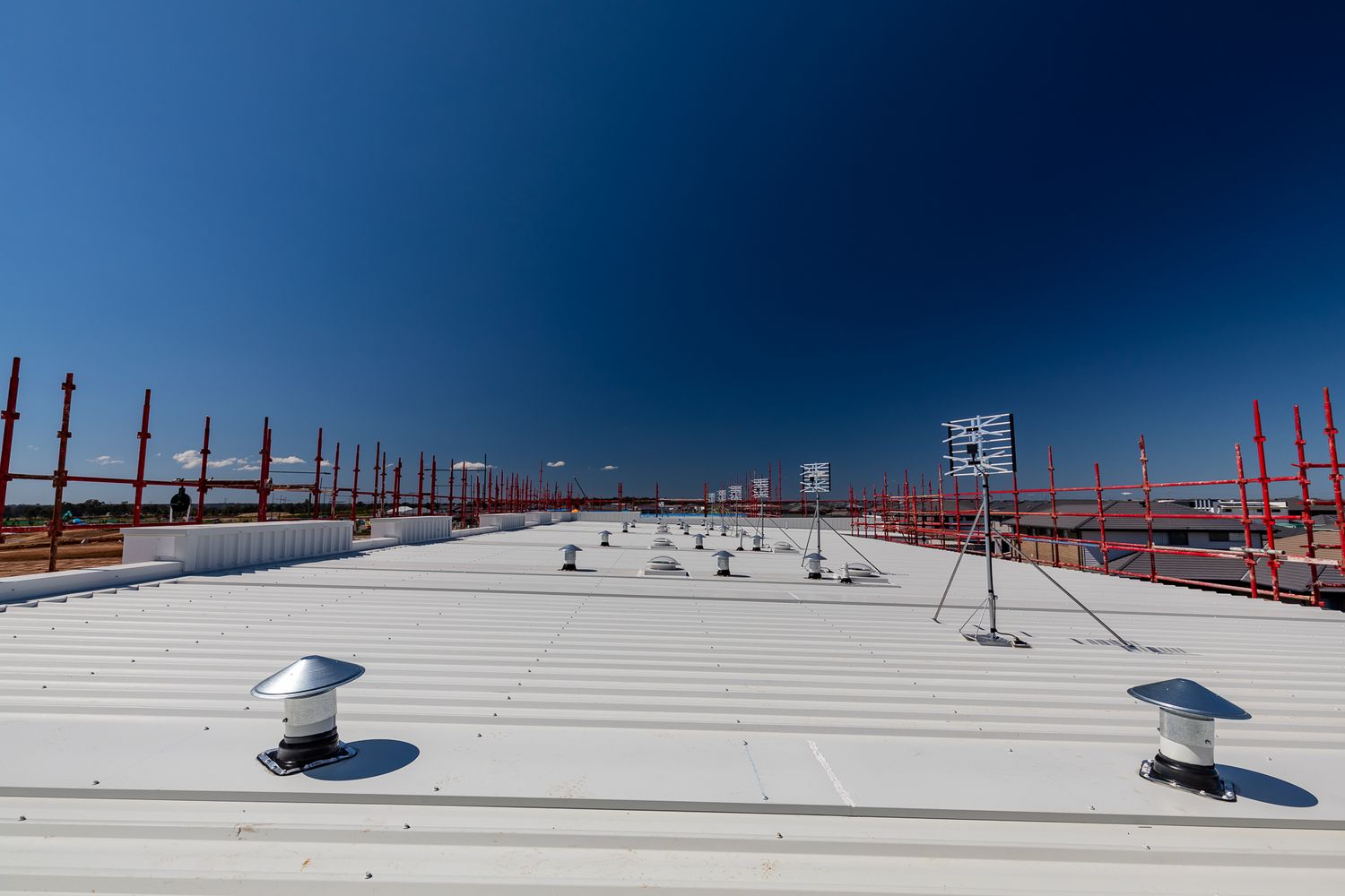 Flat metal rooftop of a new building under a deep blue sky, with roof vents, a weather sensor mast and red safety railings.