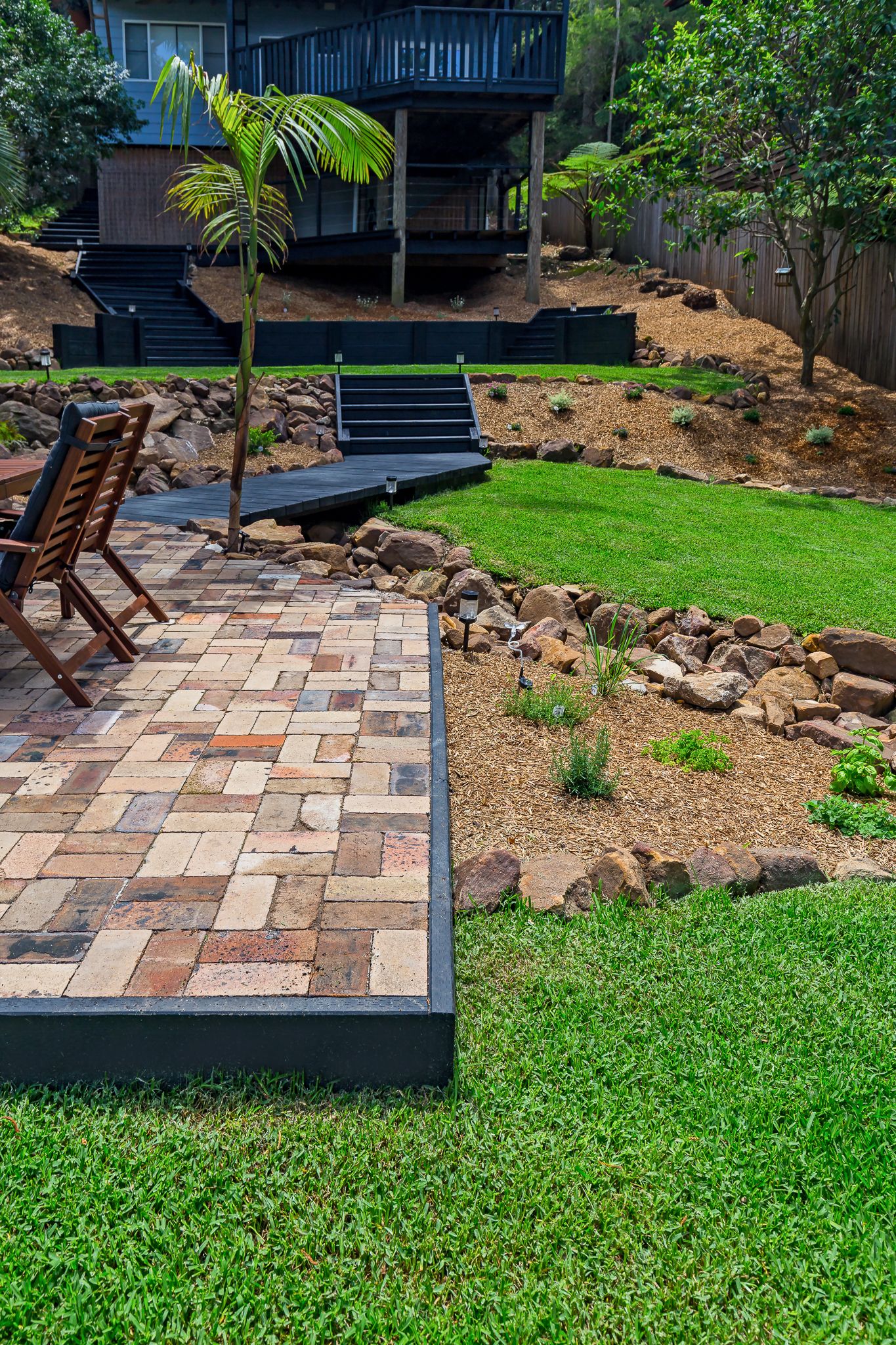 Backyard landscape with a brick paved patio, wooden deck chairs, rock garden, green lawn and a multi-level house on a hillside.