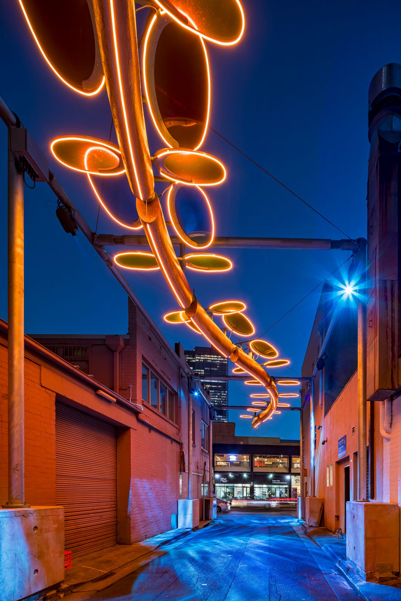 Warm orange neon oval ring light installation above a city laneway at night, with a glowing storefront and tower in the distance.