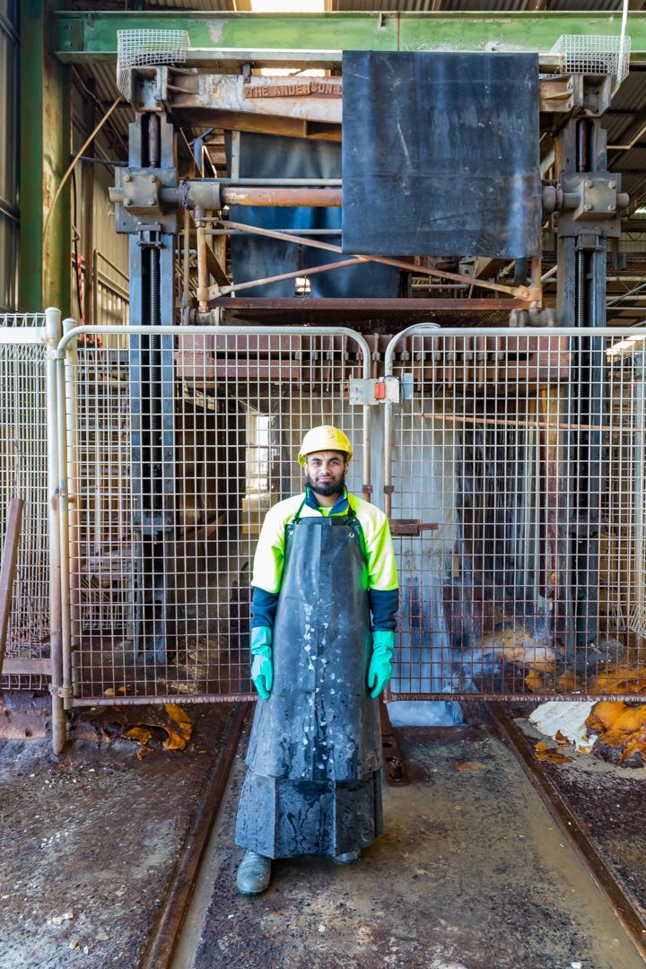 Worker in a yellow hard hat, green gloves and a rubber apron stands before an industrial machine behind a wire mesh safety fence.